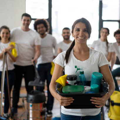 Beautiful cleaning woman with her team at an office holding a bucket with cleaning products all smiling at camera - Service concepts